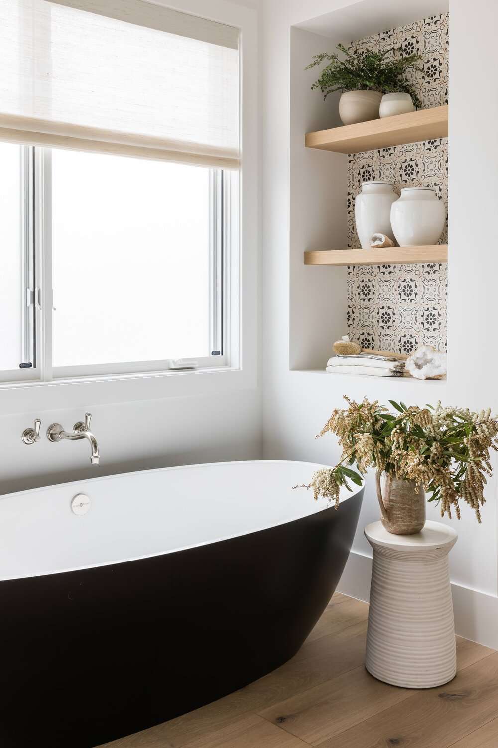 Une salle de bains avec une baignoire noire et plusieurs étagères en bois soutenues par du papier peint imprimé.